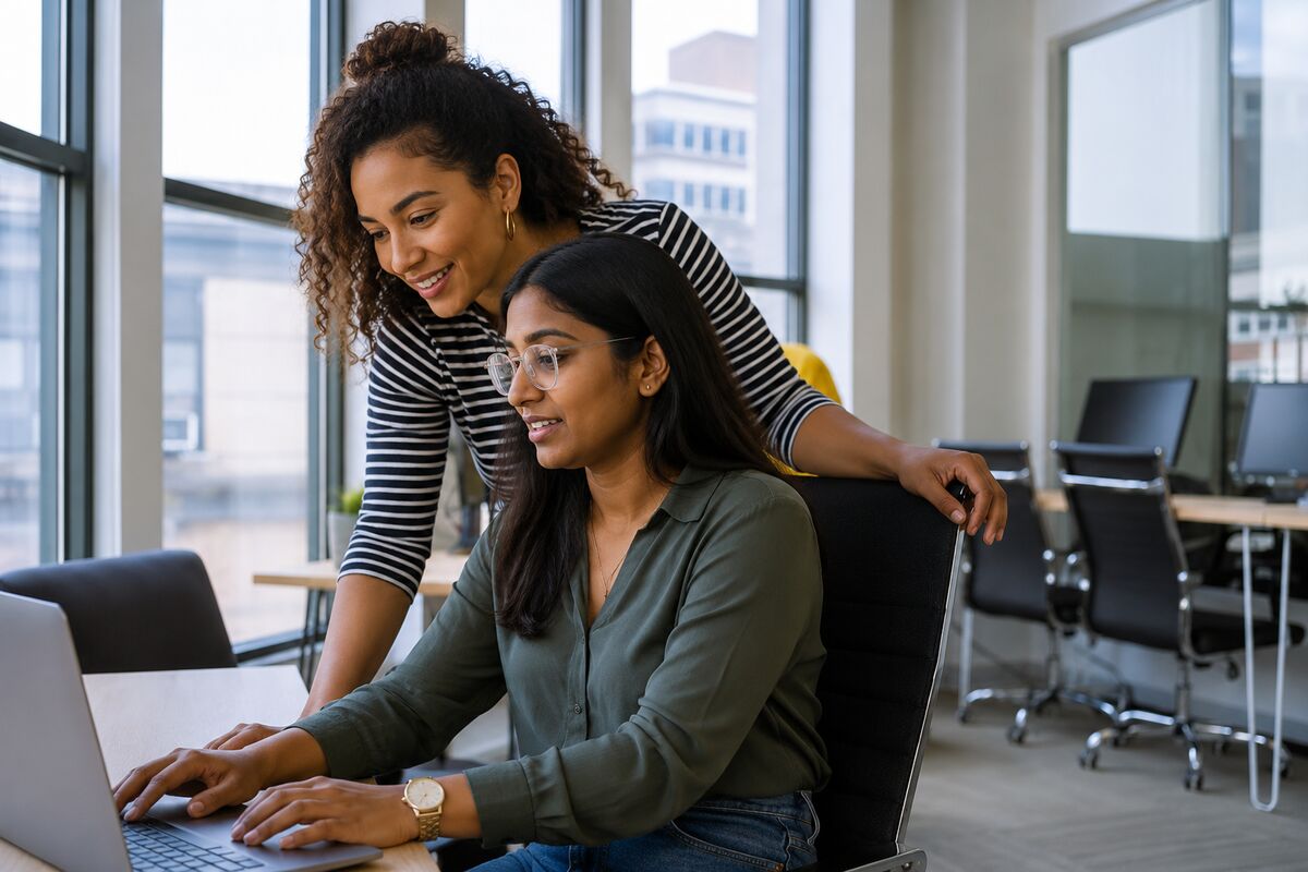 A senior colleague coaching a learner at a laptop in a professional office
