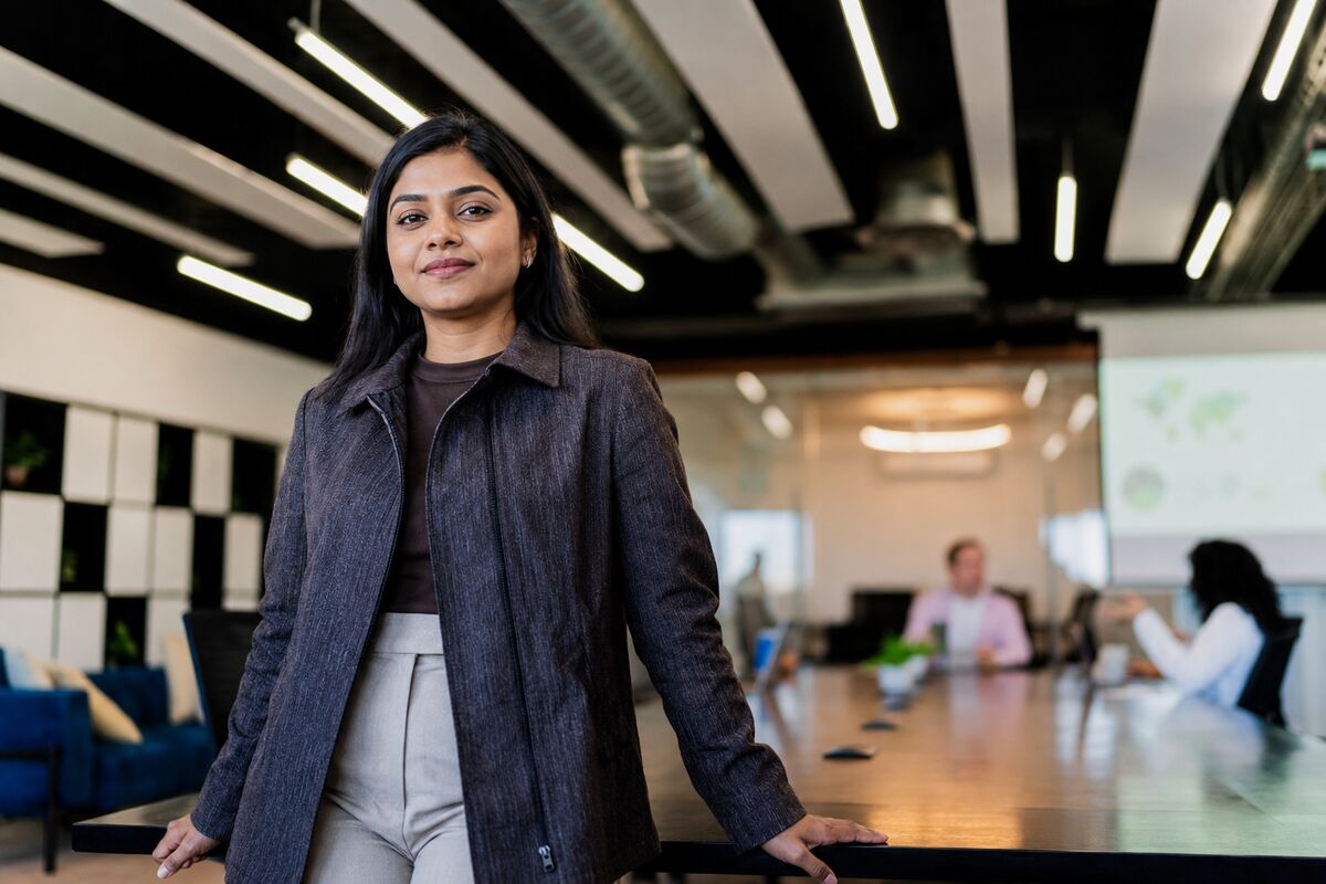 A professional standing at the head of a meeting room while colleagues work behind her